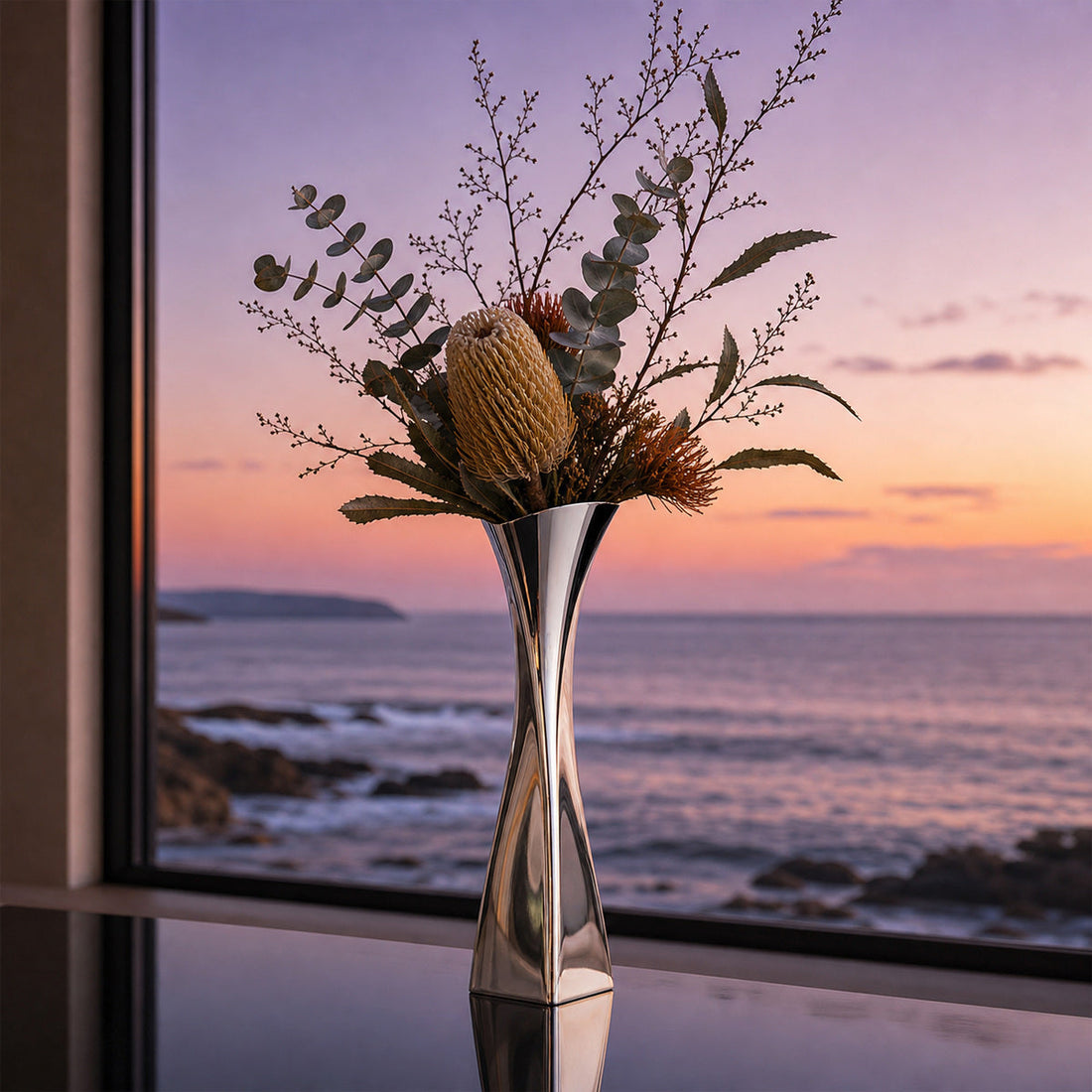 Versa Vase with winter flower arrangements with banksia and dry branches at a window view of sea at sun set.