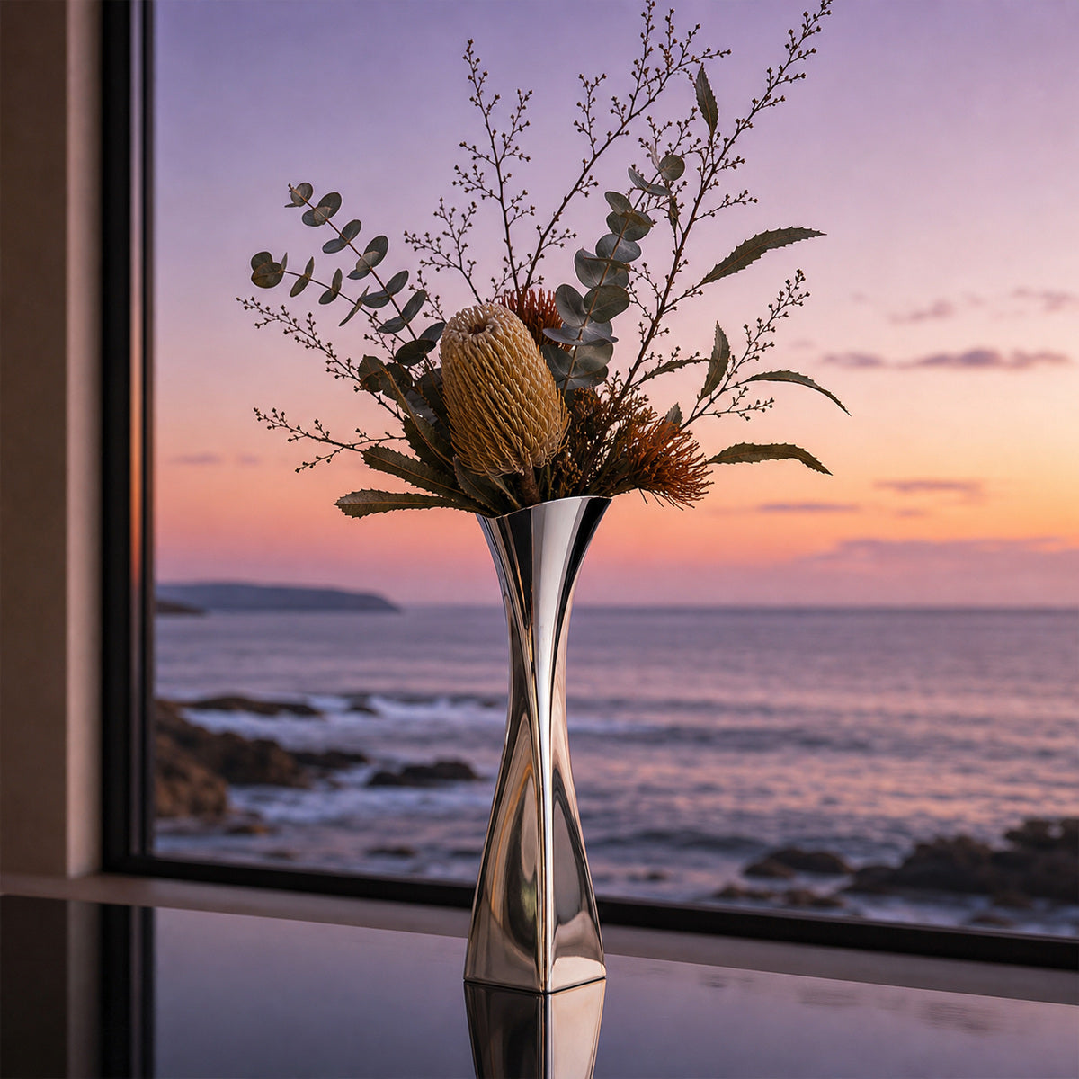 Versa Vase with winter flower arrangements with banksia and dry branches at a window view of sea at sun set.