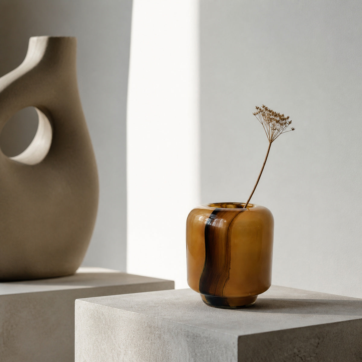 Onda Vase Boleto: a small amber-brown glossy ceramic vase with a dark vertical stripe, holding a dried flower stem, displayed on a minimalist concrete pedestal against a white wall.