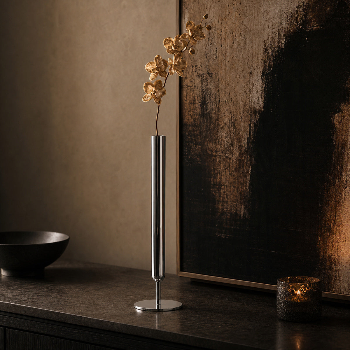 A tall, ultra-thin cylindrical metal vase, the Lumina Vase Axis, displaying a single stem of dried white orchid. The minimalist architectural  metal vase stands on a dark textured stone console in a moody, gallery-inspired room, backed by a dramatic abstract painting and a glowing votive candle.