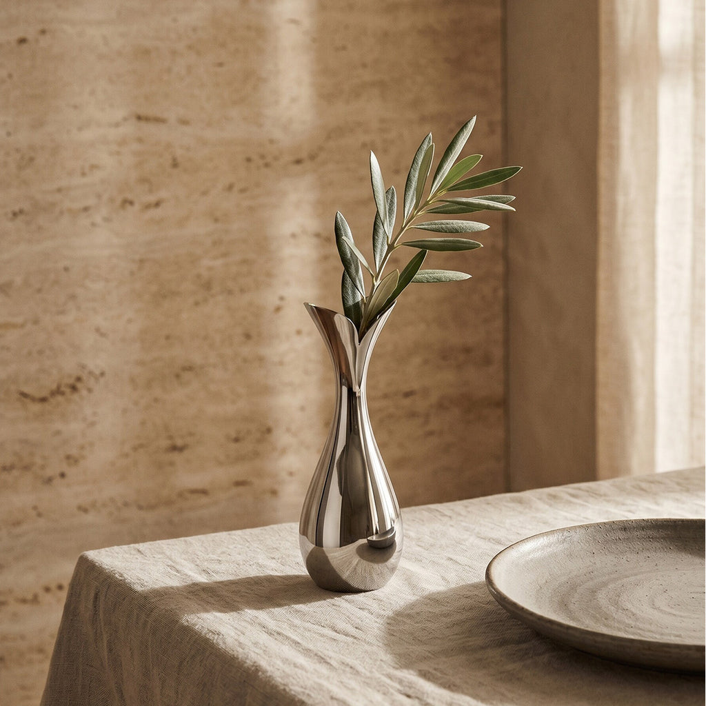 Silver stainless steel modern vase with olive tree branch on a linen lined wooden surface with a limestone wall