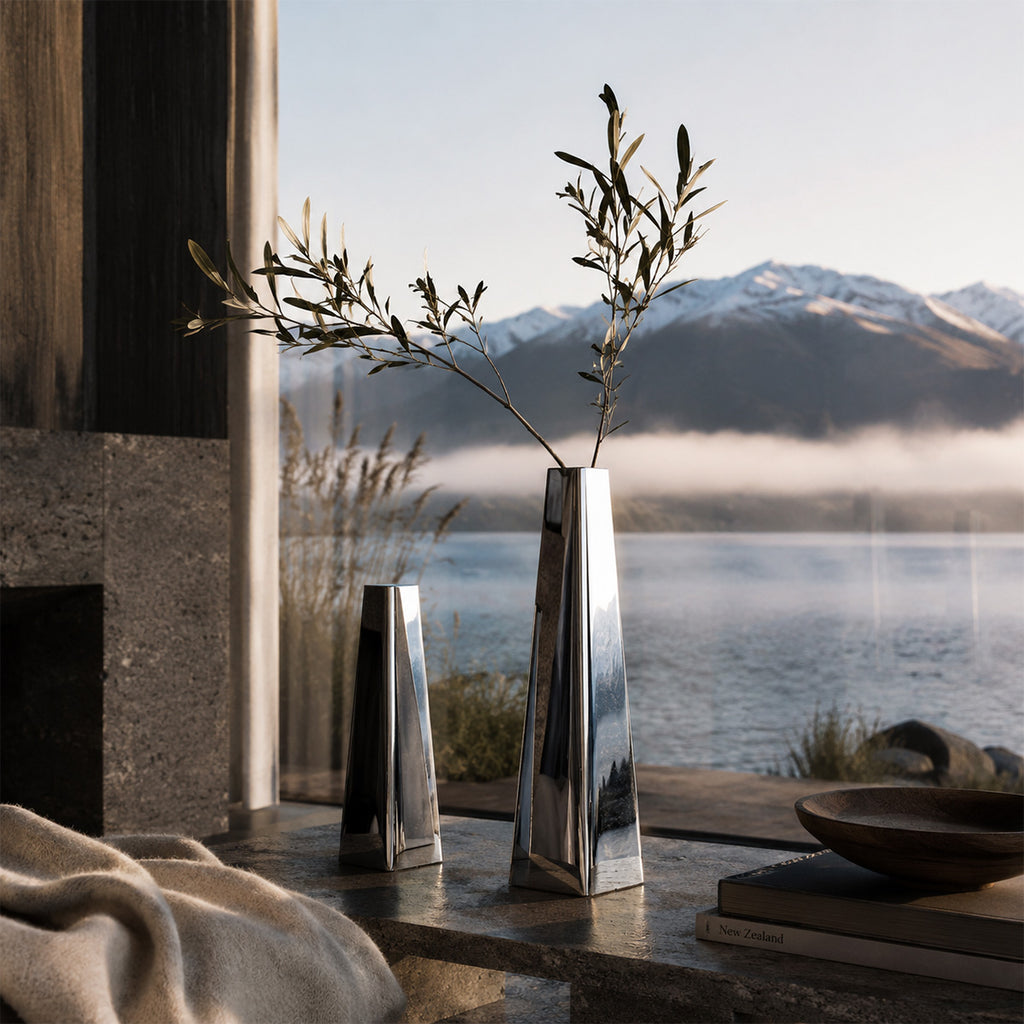 Two metallic polished stainless steel vases with a plant on a table with a scenic lakeside retreat view.