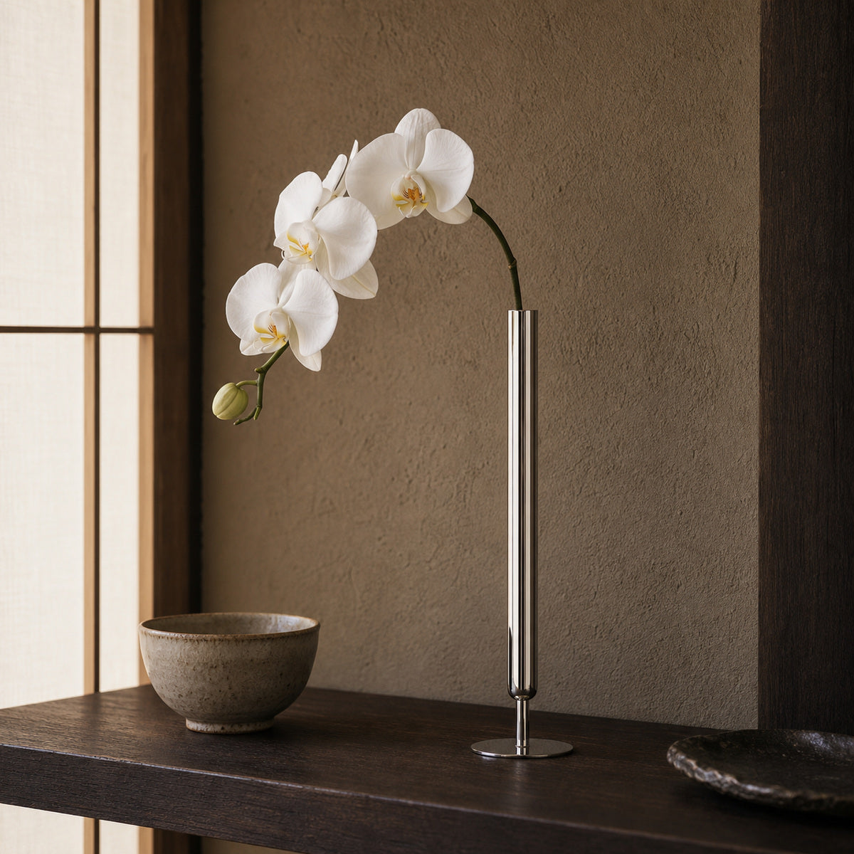 A sleek, ultra-thin cylindrical metal vase, the Lumina Vase Axis, displaying an elegant, gracefully curving fresh white orchid stem. The minimalist silver vase sits on a dark wooden shelf alongside rustic ceramic bowls, framed by an earthy textured wall and a traditional shoji screen in a serene Japandi-style setting.