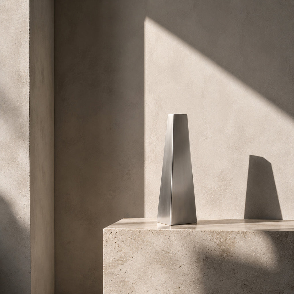 A contemporary silver geometric vase, the Lumina Vase Tactus, showcased as an art sculptural piece. The smooth metallic finish sharply contrasts with the organic concrete block it sits on, illuminated by high-contrast natural light in a minimalist interior.