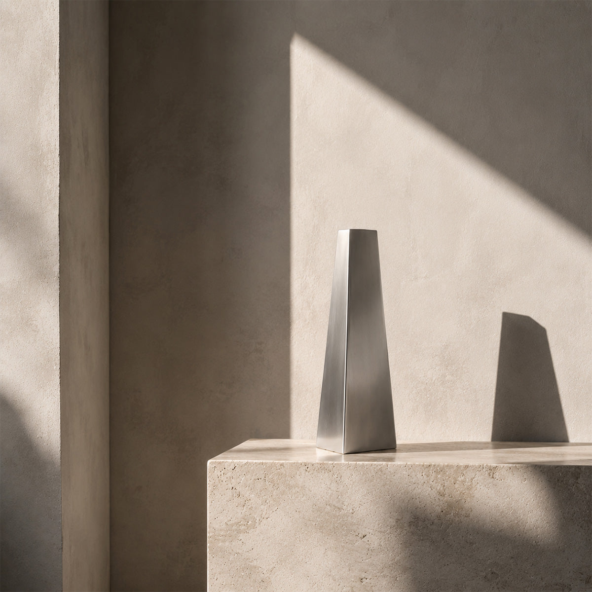 A contemporary silver geometric vase, the Lumina Vase Tactus, showcased as an art sculptural piece. The smooth metallic finish sharply contrasts with the organic concrete block it sits on, illuminated by high-contrast natural light in a minimalist interior.