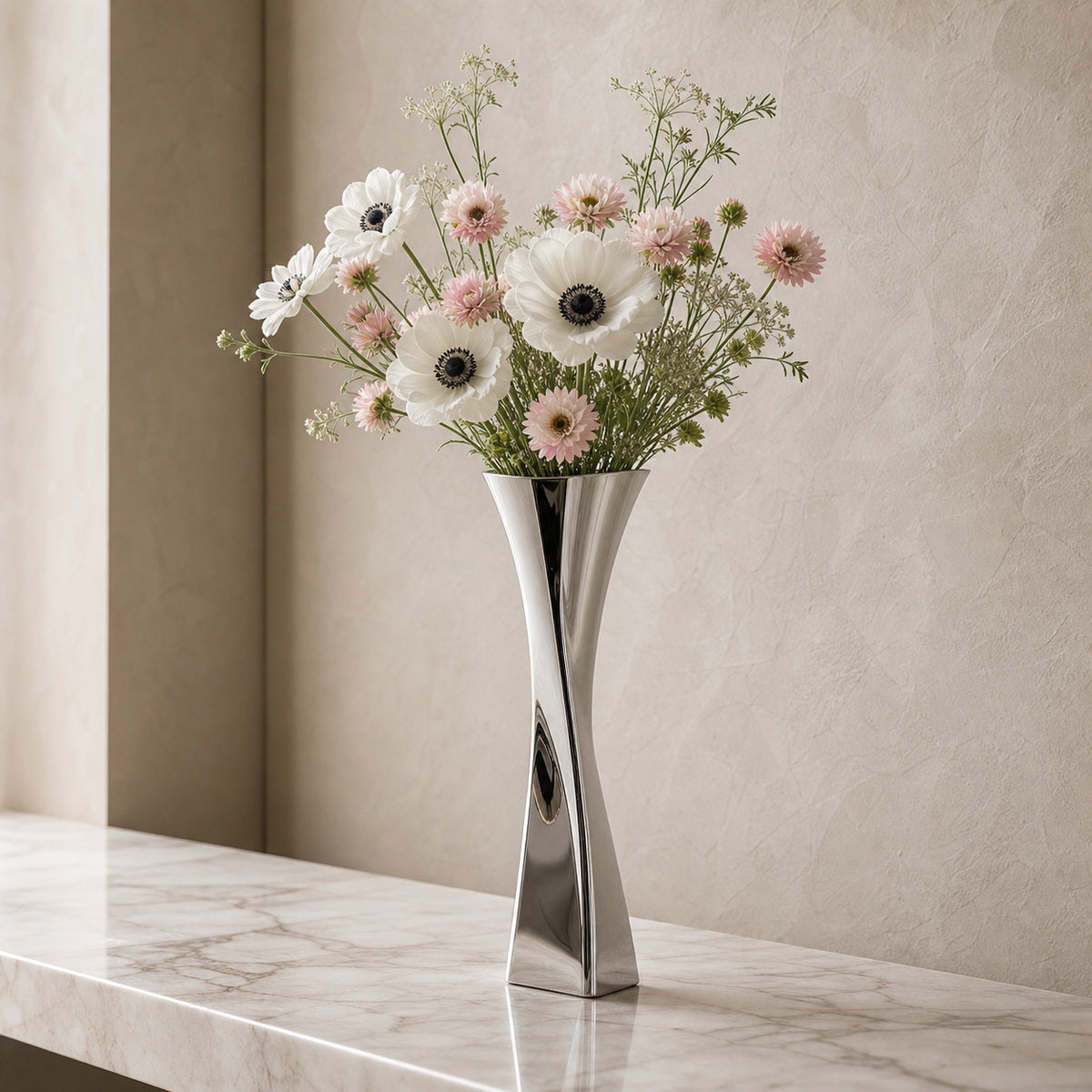 A polished silver metal vase, the Lumina Vase Versa V, displaying a romantic floral arrangement of white anemones and pastel pink blooms. The twisted geometric vase sits on a luxurious marble ledge against a warm textured plaster wall in an elegant, bright setting.