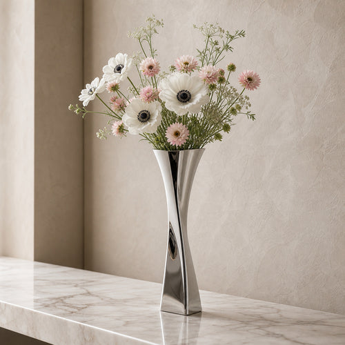 A polished silver metal vase, the Lumina Vase Versa V, displaying a romantic floral arrangement of white anemones and pastel pink blooms. The twisted geometric vase sits on a luxurious marble ledge against a warm textured plaster wall in an elegant, bright setting.