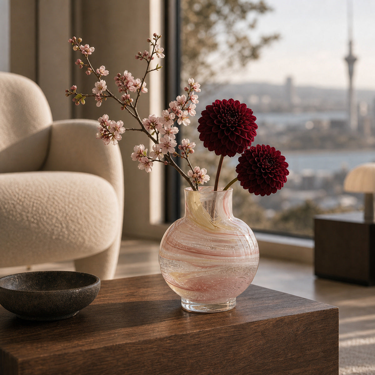 Decorative pink hand made glass vase with flowers on a wooden coffee table in lounge room with a city view.