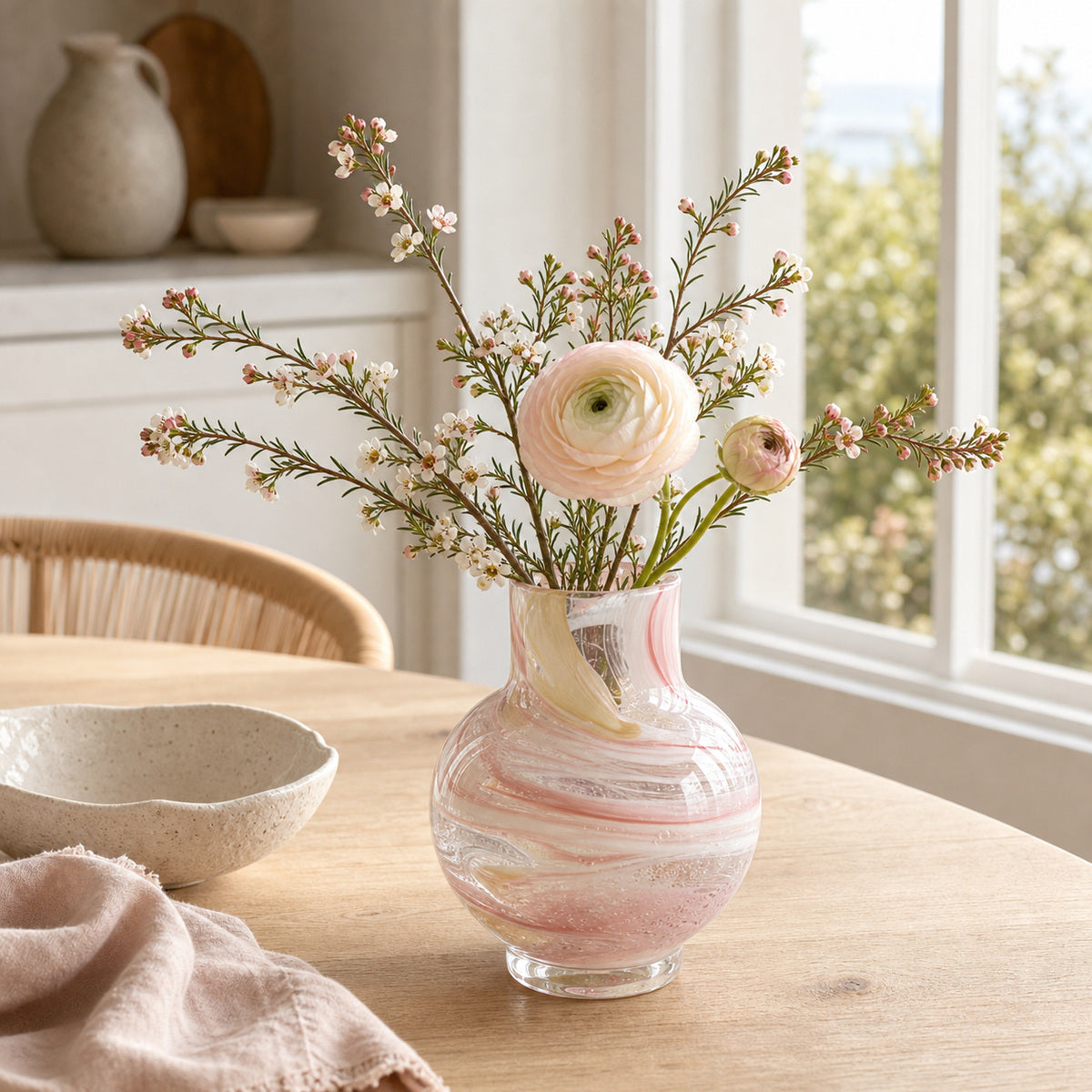 Pink glass vase with fresh flowers on a wooden dining table near a window