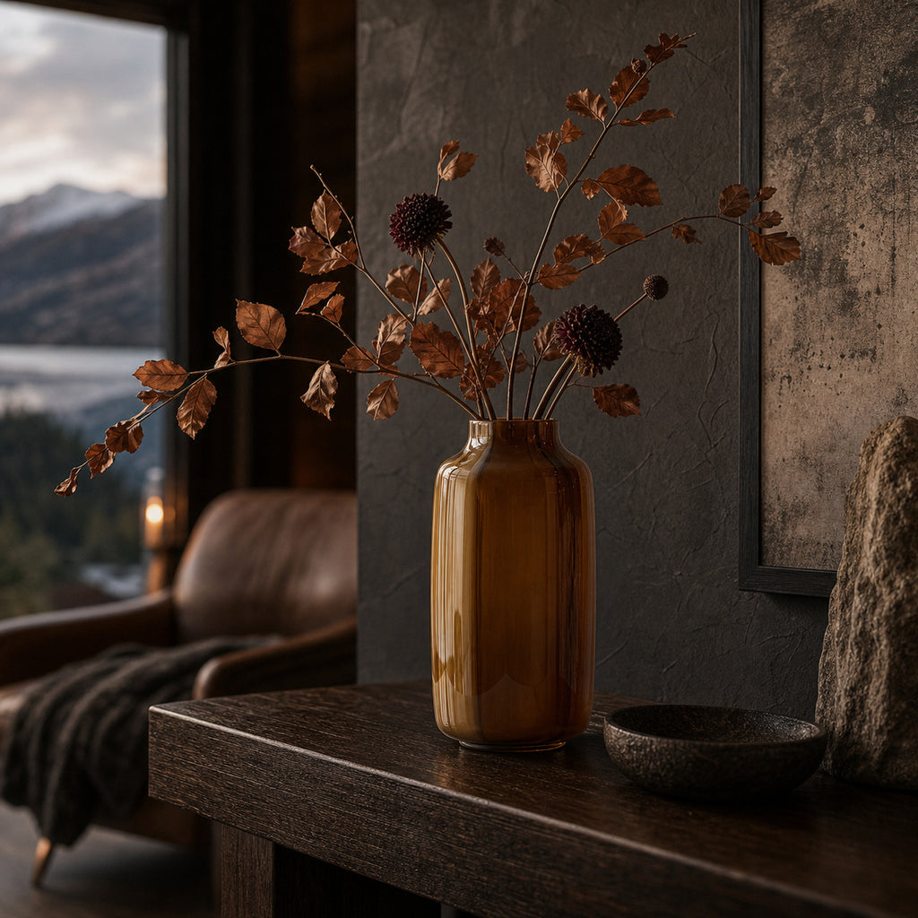 Brown opaque modern glass vase with dried autum leaves and flowers on a dark wooden table in a room with a window view of mountains and lakes.