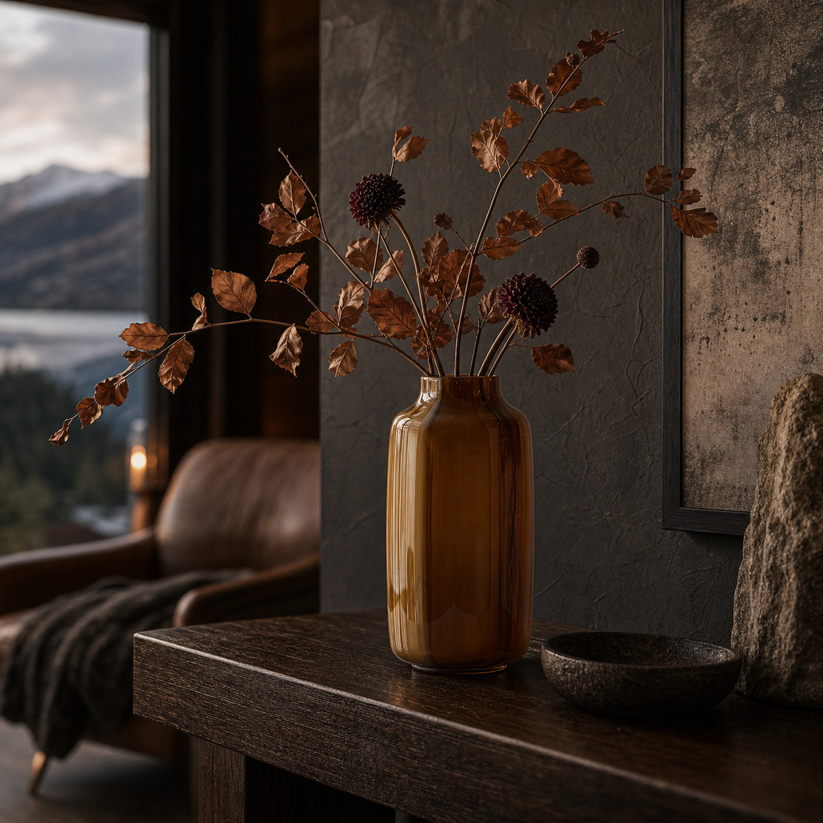 Brown opaque modern glass vase with dried autum leaves and flowers on a dark wooden table in a room with a window view of mountains and lakes.
