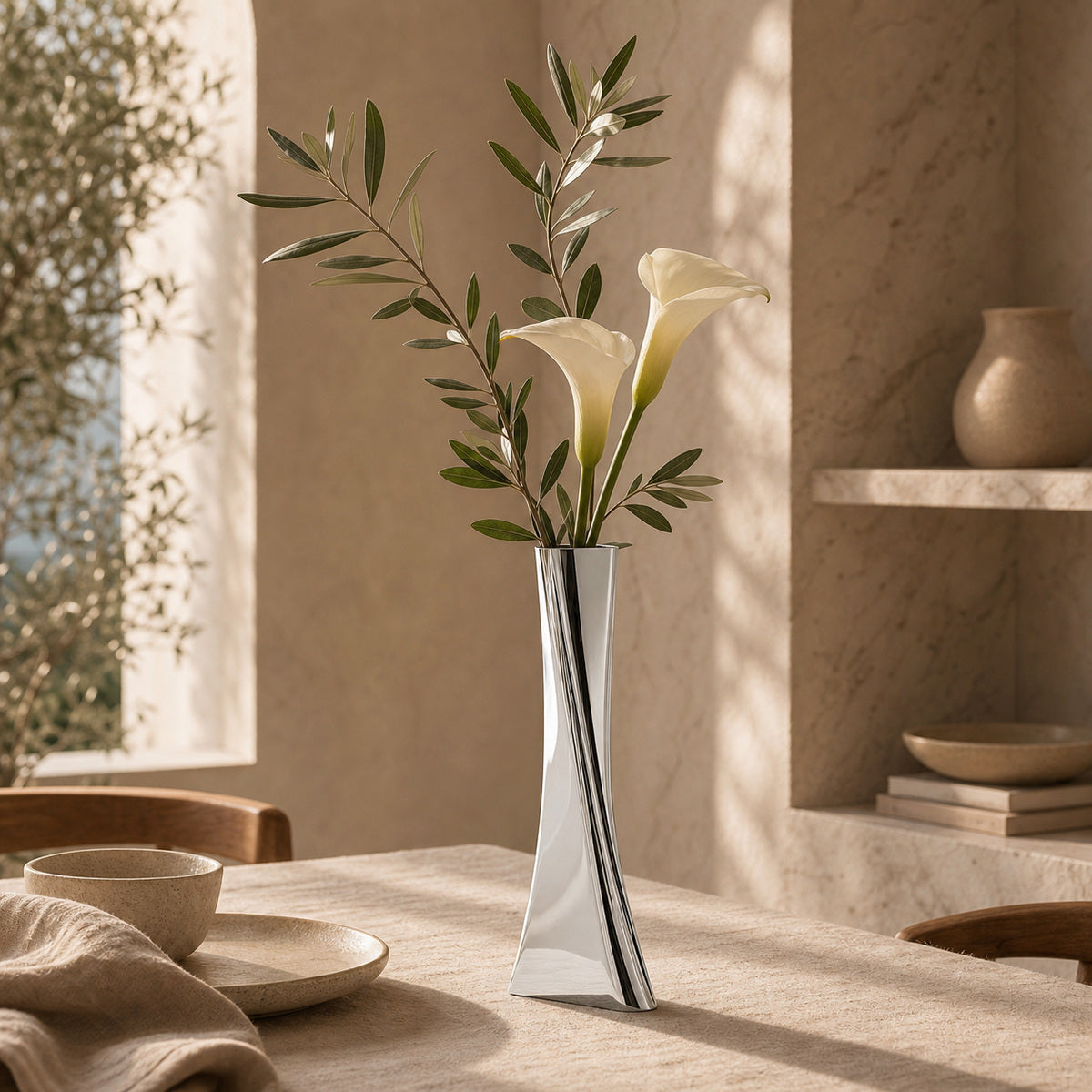 Silver vase with white flowers and green leaves on a table in a softly lit room.