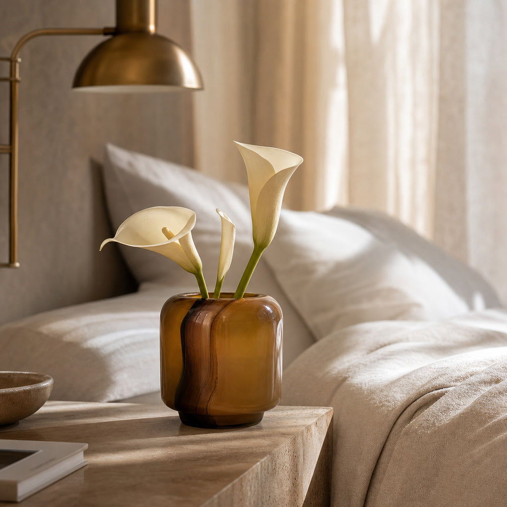 Onda Vase Boleto: an amber glass vase with rounded geometric edges holding three cream calla lilies, placed on a light wood bedside table in a softly lit bedroom with beige bedding, pillows, and a brass wall lamp in the background.