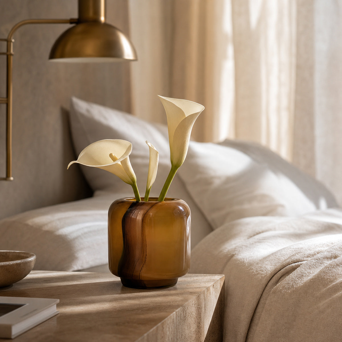 Onda Vase Boleto: an amber glass vase with rounded geometric edges holding three cream calla lilies, placed on a light wood bedside table in a softly lit bedroom with beige bedding, pillows, and a brass wall lamp in the background.