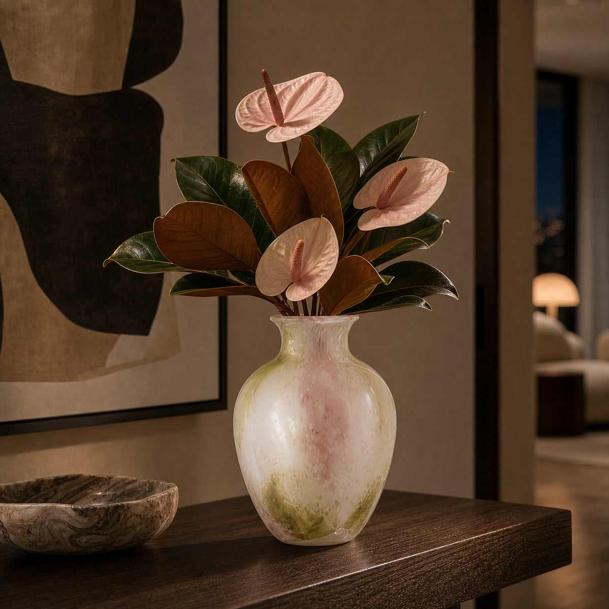 Pastel pink and green glass vase - Onda Vase Petal displaying pale pink anthuriums and dark magnolia leaves. The vase is positioned on a dark wood console table next to a marbled stone bowl in an elegant, moody hallway leading to the lounge. The sophisticated setting features a contemporary abstract painting and a softly lit interior.