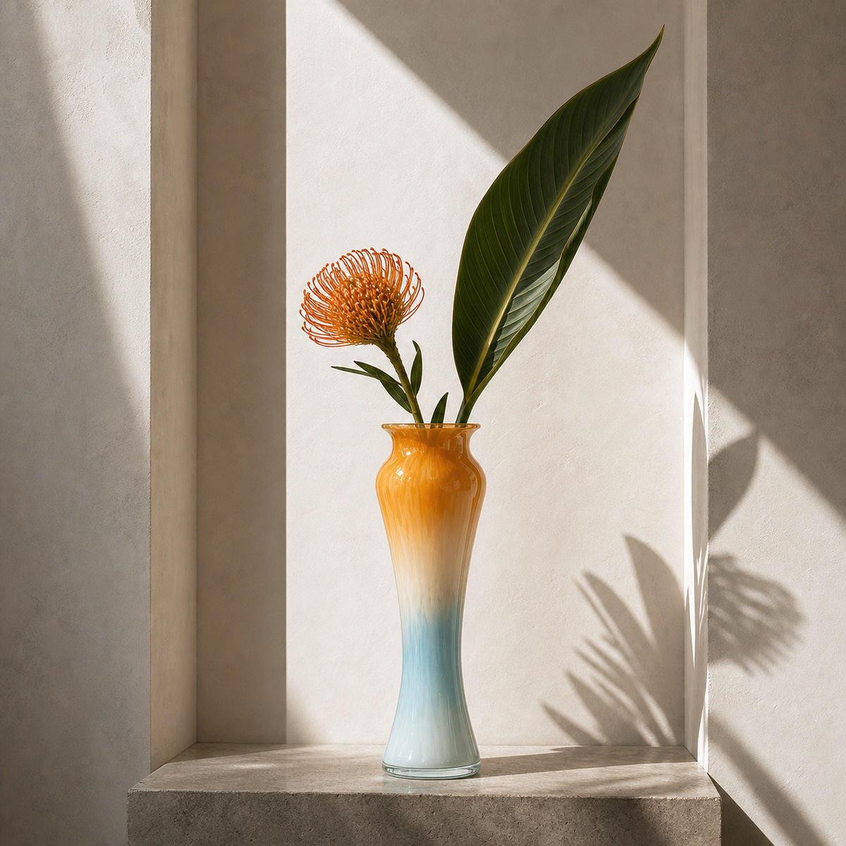 Gradient Glass Vase - Onda Vase Solis in an orange and blue gradient, displaying an orange pincushion protea and a tropical leaf on a concrete plinth in a sunlit wall niche.