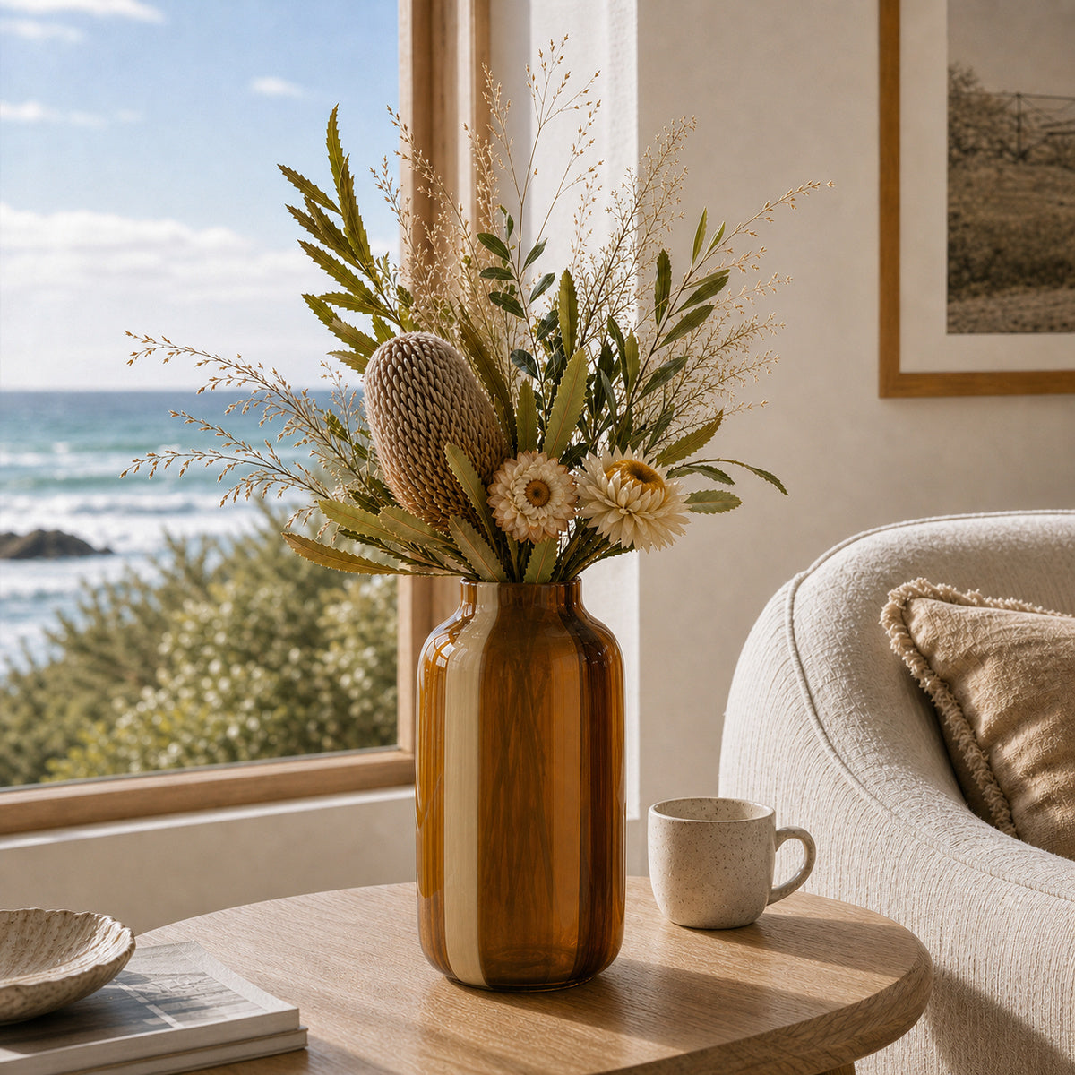 Striped glass vase - Onda Vase Toffane in amber and cream, displaying dried native flowers including a prominent Banksia and strawflowers. The vase is styled on a round wooden coffee table next to a cream textured fabric armchair in a bright, cozy living room overlooking the ocean.