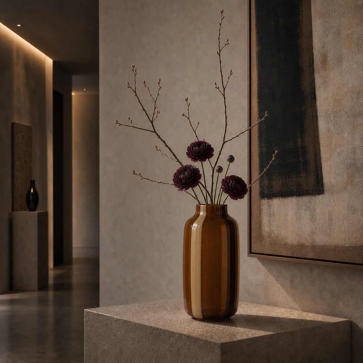 A detailed interior view featuring the two-tone (caramel and dark brown) striped glass Onda Vase Toffane. The vase is centered on a rough, textured stone pedestal in a minimalist modern living space. It holds a composition of delicate bare branches and several deep burgundy-red ranunculus flowers. The setting includes soft, warm, recessed LED ceiling lighting, a dark polished floor, and a large abstract painting with a dark vertical form on the wall. The atmosphere is calm and luxurious.