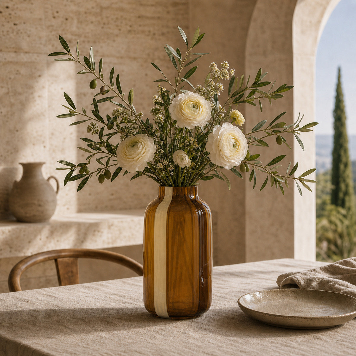 Striped glass vase - Onda Vase Toffane in caramel and cream, styled with white ranunculus and fresh olive branches. The vase sits on a linen-draped table in a sunlit Mediterranean-style room with stone archways.