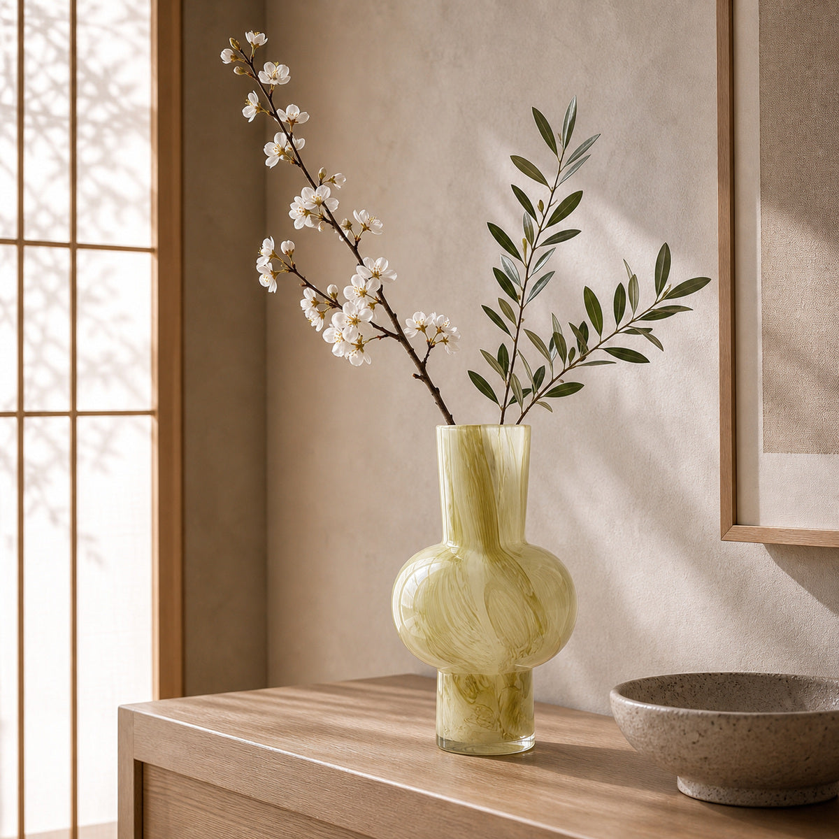 Green marbled glass vase - Onda vase verde displaying a white blossom branch and green foliage. The vase sits on a light wood console next to a textured ceramic bowl in a serene Zen-inspired interior. Sunlight filters through a traditional wooden lattice screen, casting elegant shadows on the warm plaster wall.