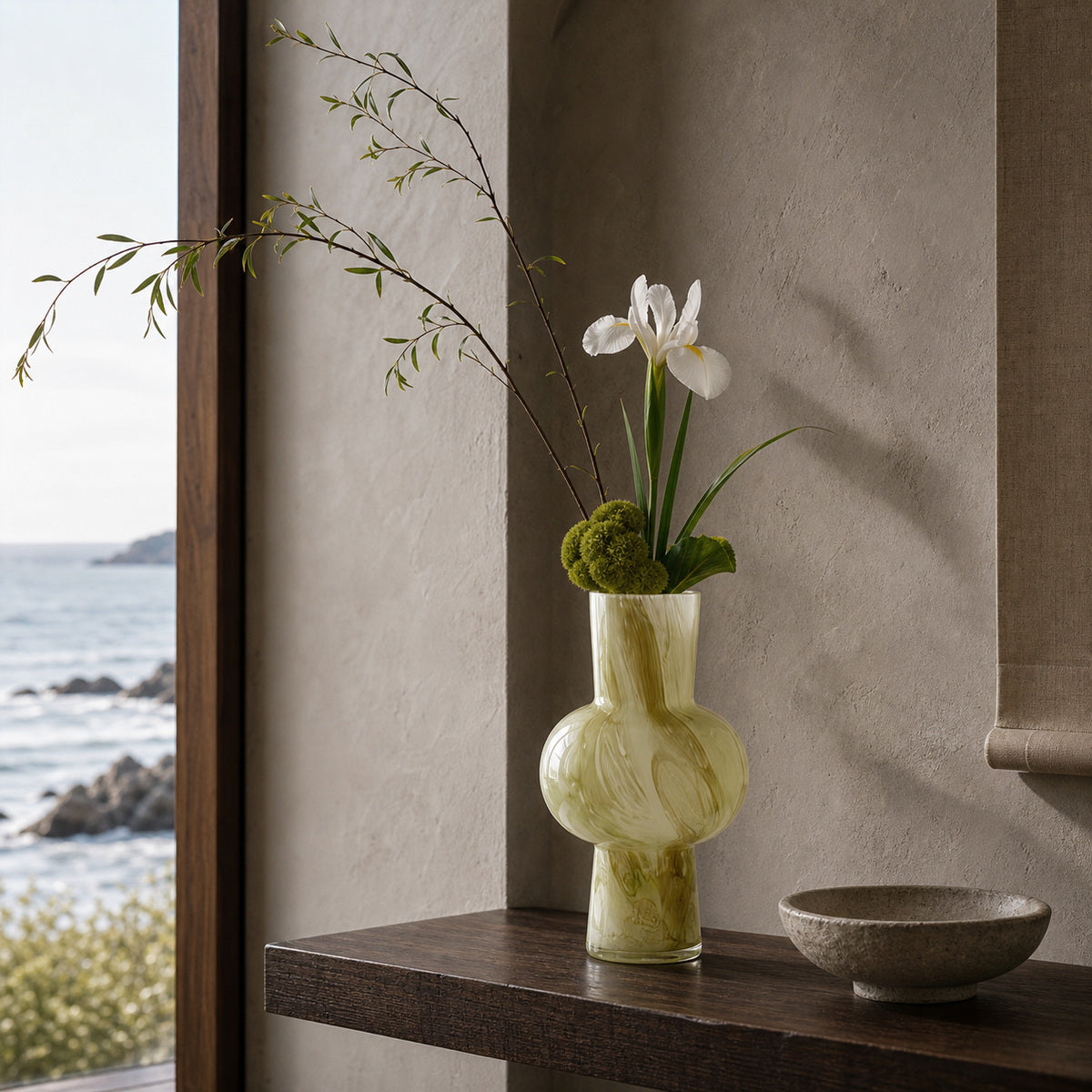 Onda Vase Verde featuring a green swirl pattern, styled with an elegant minimalist floral arrangement on a dark wooden console. The Coastal Zen setting highlights textured plaster walls, natural shadows, and a window framing the sea.